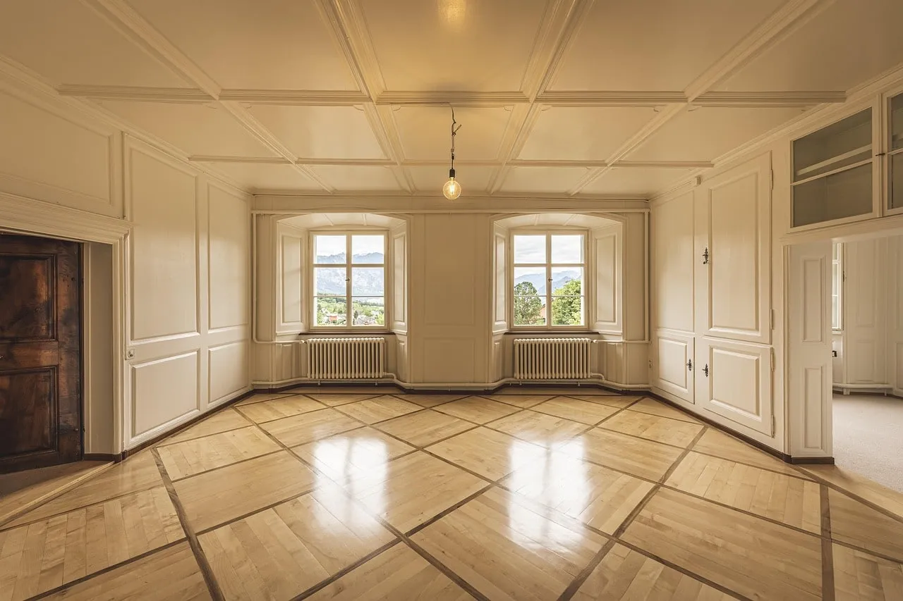 Ein leerer Altbauraum mit Parkettboden, an der Decke hängt eine nackte Glühbirne zwischen zwei Fenstern