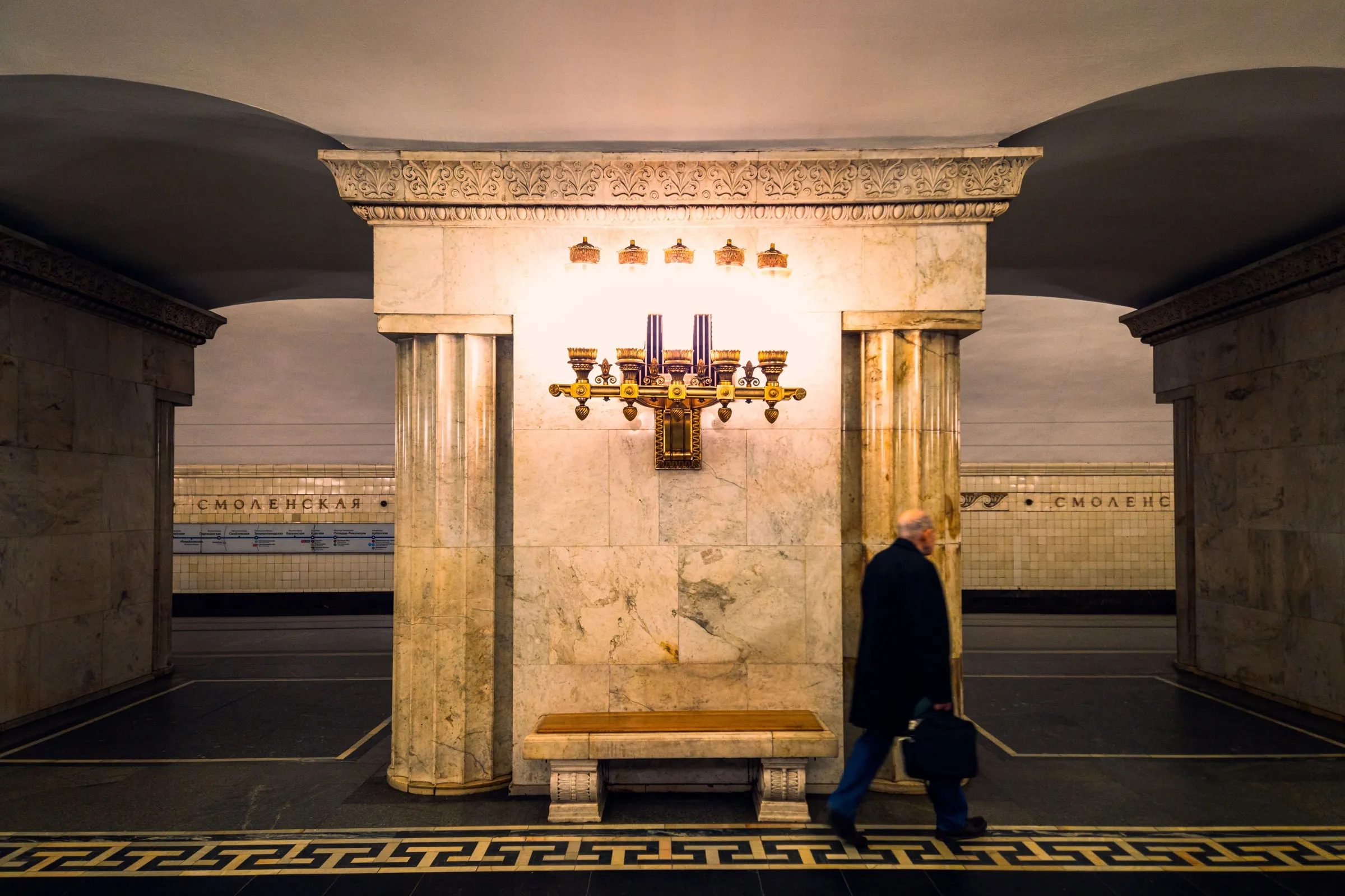 Die Marmorhalle der Moskauer Metrostation Smolenskaja, an der Wand ein bronzener Leuchter, davor geht ein Mann vorbei
