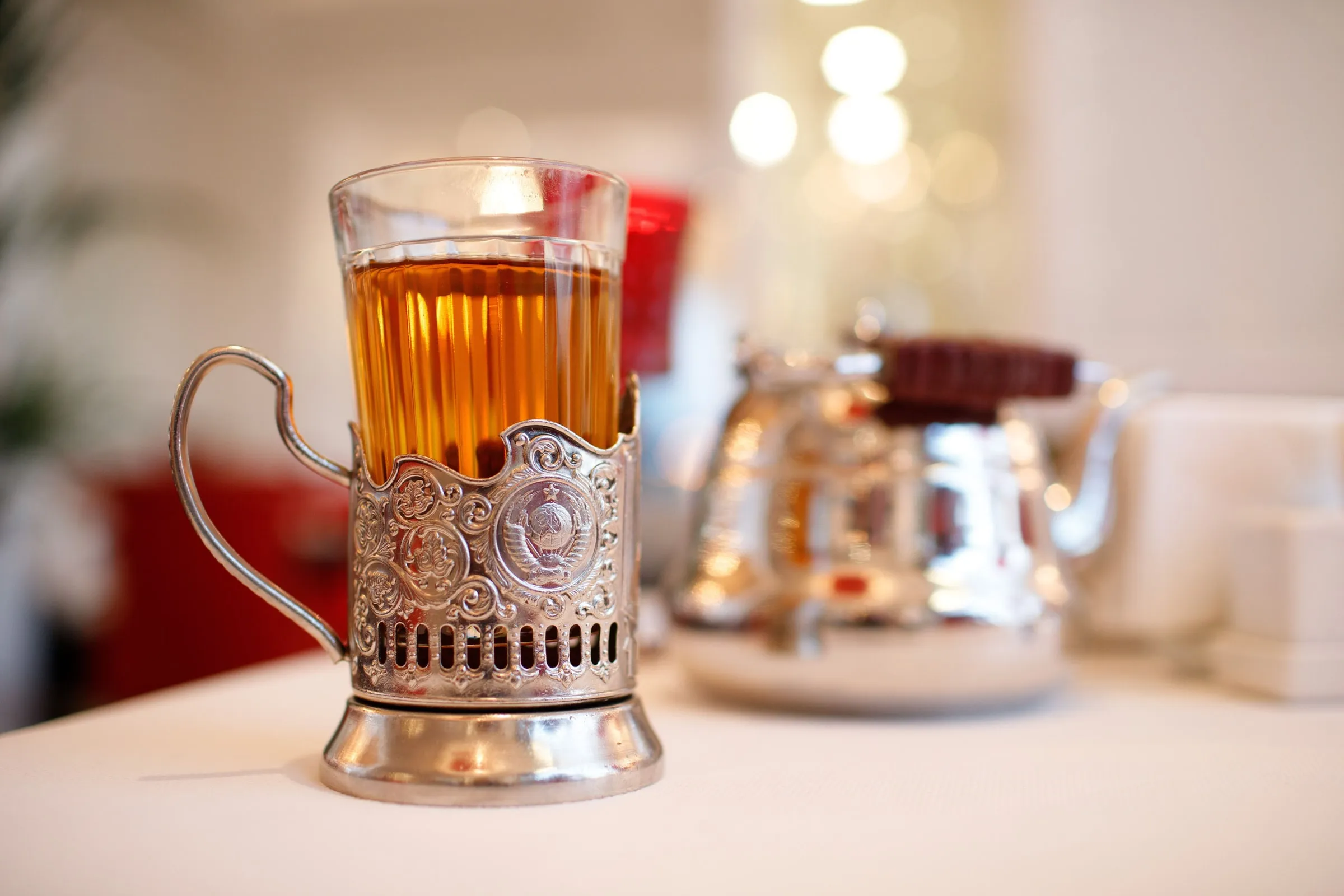 Ein Glas Tee in einem verzierten Metall-Glashalter steht auf einem weißen Tisch, dahinter unscharf eine silberne Teekanne