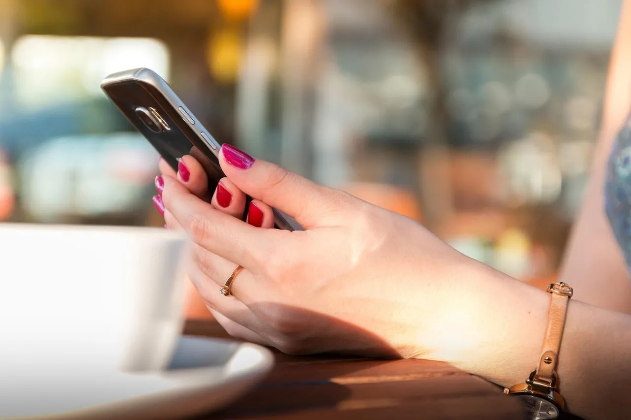 Zwei Hände halten ein Smartphone über einem Cafétisch, im Hintergrund unscharfe Lichter