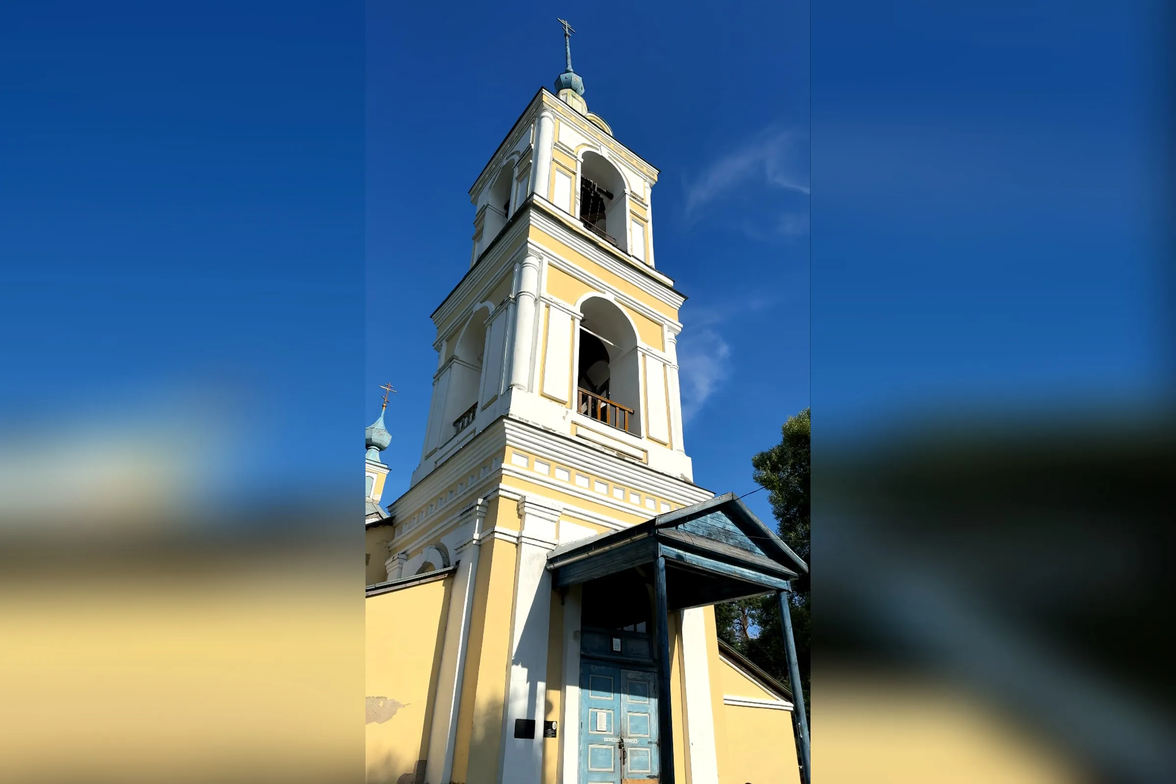 Der gelb-weiße Glockenturm der Troizki-Kirche in Troizkoje an der Ustje, davor das blaue hölzerne Vordach über dem Eingang