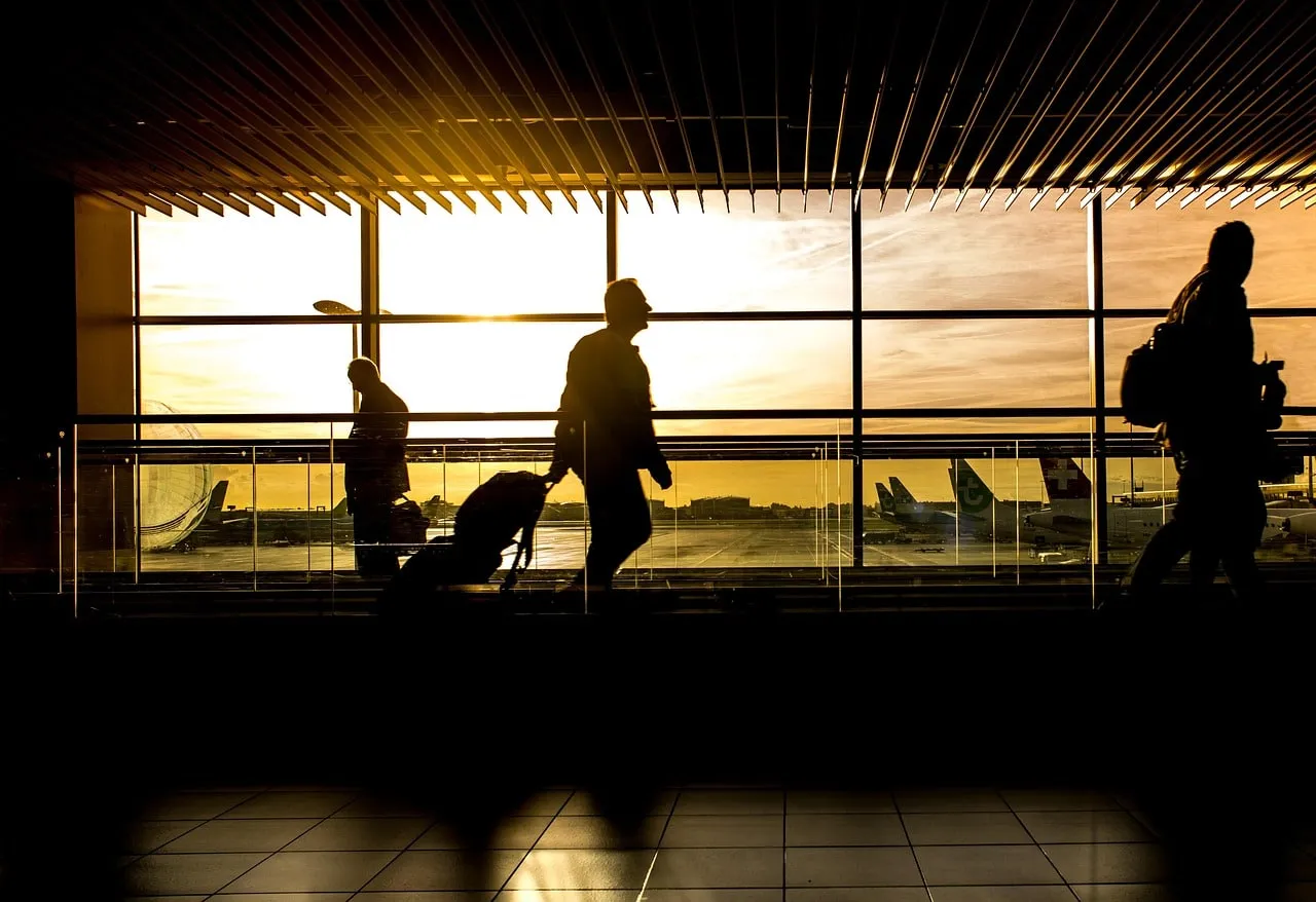 Reisende mit Gepäck als Silhouetten vor der Fensterfront eines Flughafenterminals, dahinter Flugzeuge im Gegenlicht der tiefstehenden Sonne